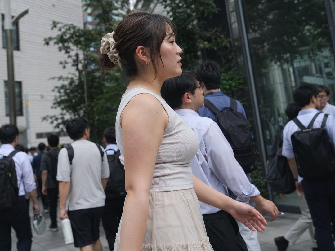 A young East Asian woman with her hair tied up walks on a busy city street during the daytime, wearing a light-colored tank top and skirt.