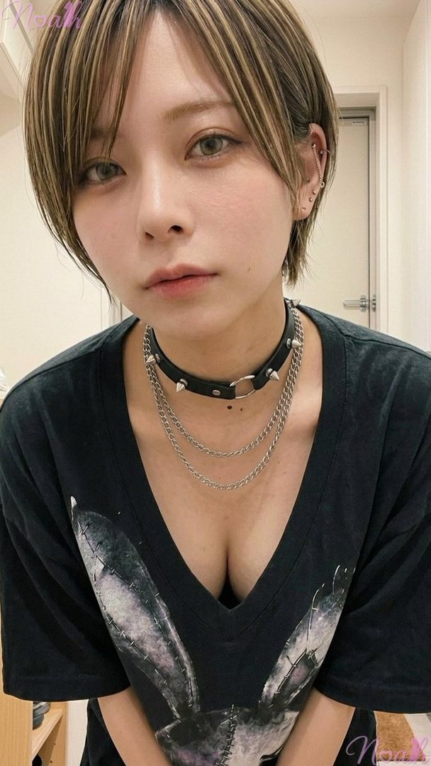 A young woman with short brown hair and blonde highlights wears a black v-neck t-shirt and a spiked choker with chains, looking directly at the camera.