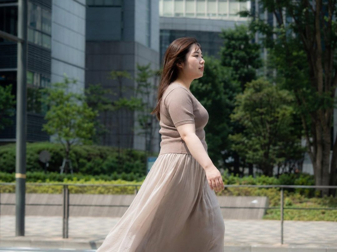 A young woman with long brown hair walks down a city street, wearing a beige knit top and a long beige skirt.