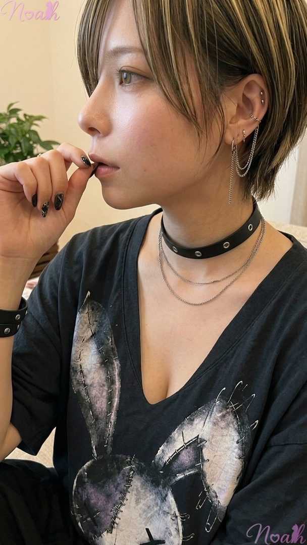 A woman with short, highlighted hair and multiple piercings wears a black t-shirt with a bunny design, a studded choker, and chain earrings, with her hand near her mouth.
