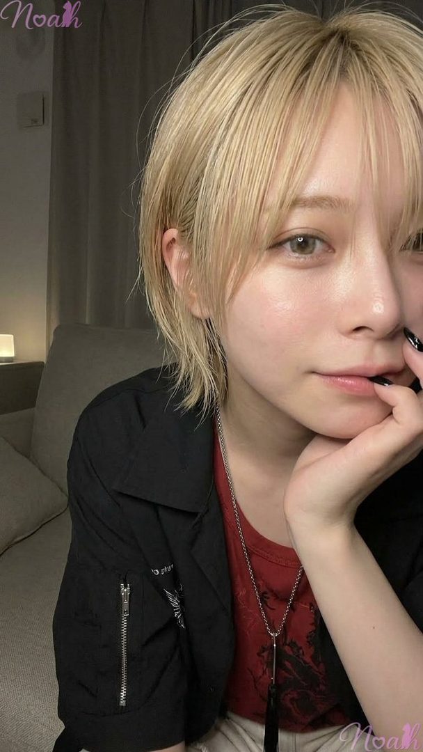 A person with short blonde hair and light eyes, wearing a black jacket over a red top, poses indoors with their hand to their chin.