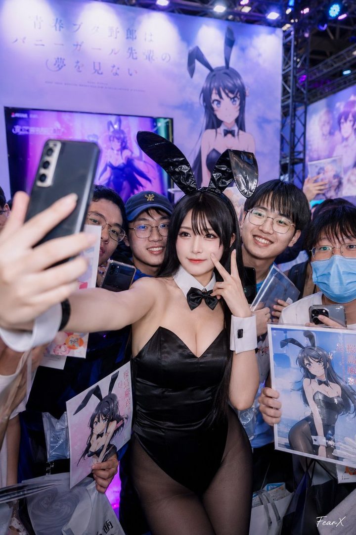 A cosplayer dressed as a bunny girl takes a selfie with fans at an event, holding up a phone and posing with a peace sign.