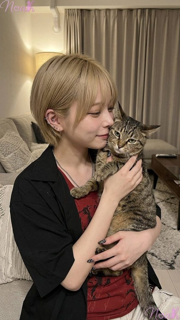 A person with short blonde hair and multiple ear piercings is affectionately holding a tabby cat indoors.