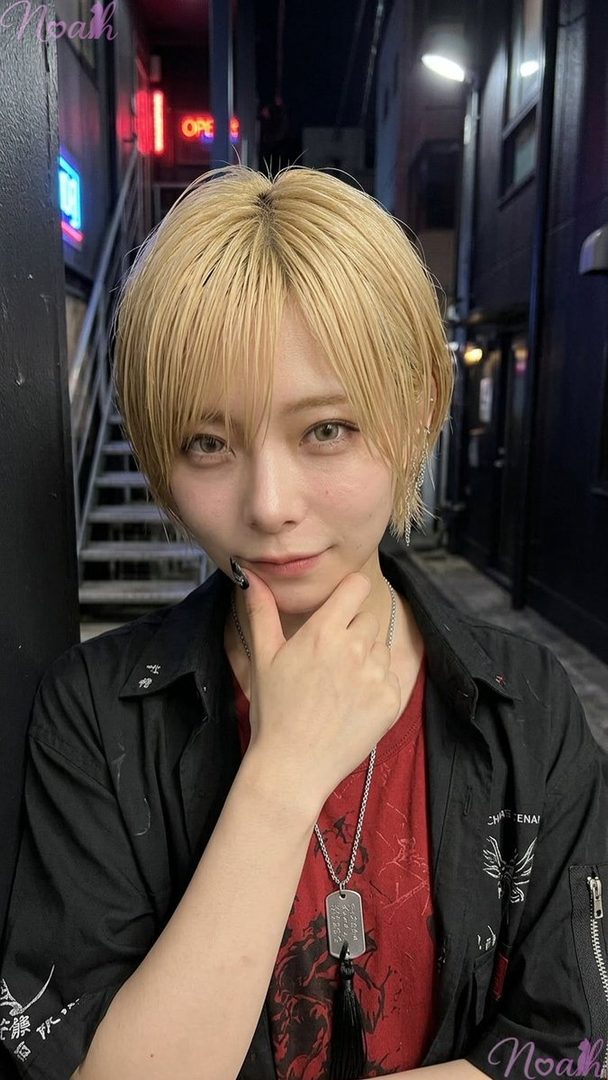 A person with short blonde hair and a black jacket over a red t-shirt poses thoughtfully in an urban alley at night.