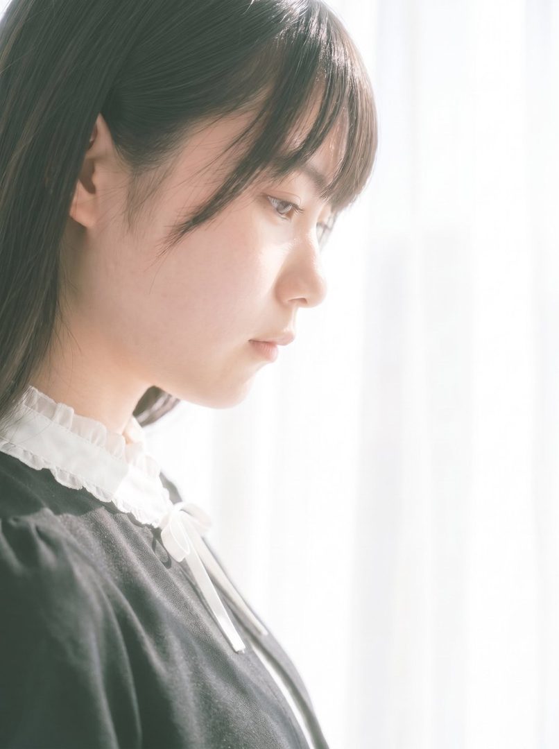 A young East Asian woman with long black hair and bangs, wearing a black dress with a white ruffled collar, looks down thoughtfully in soft natural light.