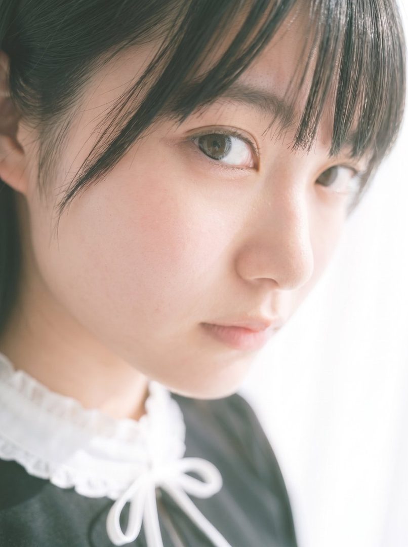 A close-up portrait of a young East Asian woman with black hair and bangs, wearing a black top with a white ruffled collar and bow, looking directly at the camera with a serene expression.