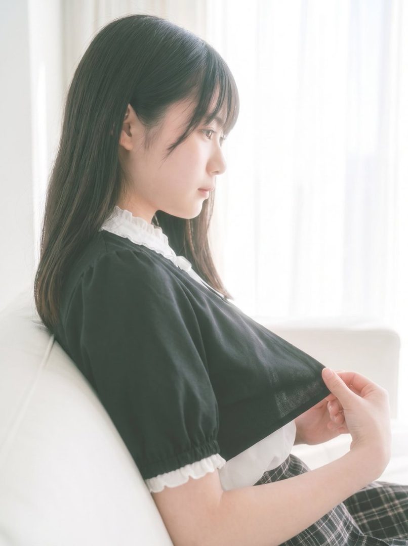 A young East Asian woman with long black hair, wearing a black dress with white lace trim, sits indoors by a window, looking thoughtfully to the side.
