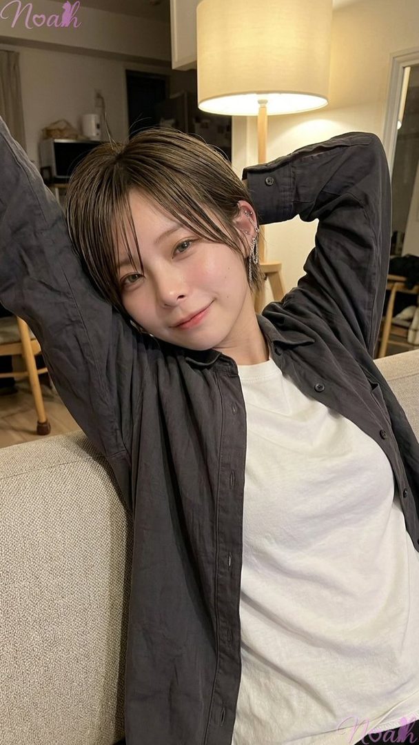 A young woman with short brown hair and bangs, wearing a dark shirt over a white t-shirt, is relaxing on a sofa indoors, looking at the camera with a gentle smile.