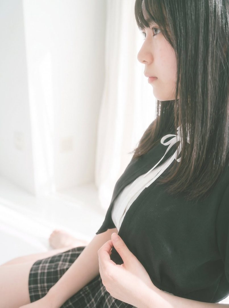 A young East Asian woman with long black hair, wearing a black cardigan over a white top and a plaid skirt, sits indoors near a window with soft light.