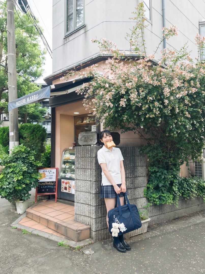 A young woman in a school uniform stands outside a patisserie, holding a piece of toast in her mouth.