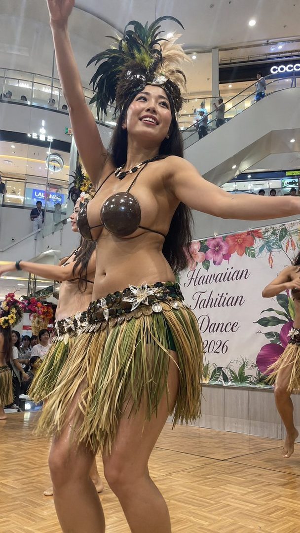 A woman in traditional Hawaiian attire, including a grass skirt and a shell top, performs a Tahitian dance on a wooden stage indoors.
