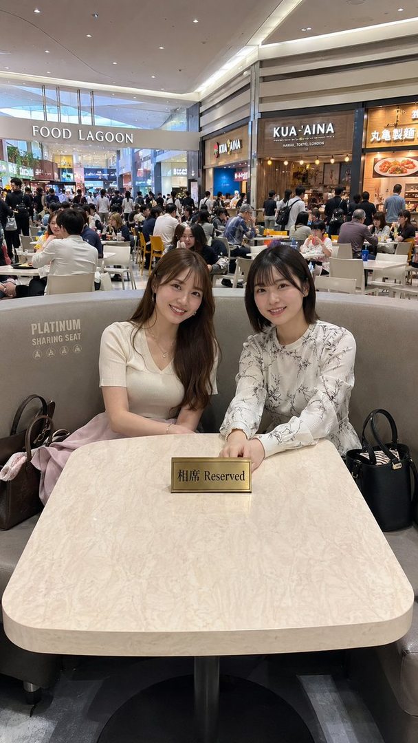 Two young East Asian women smiling while sitting at a table in a busy food court.