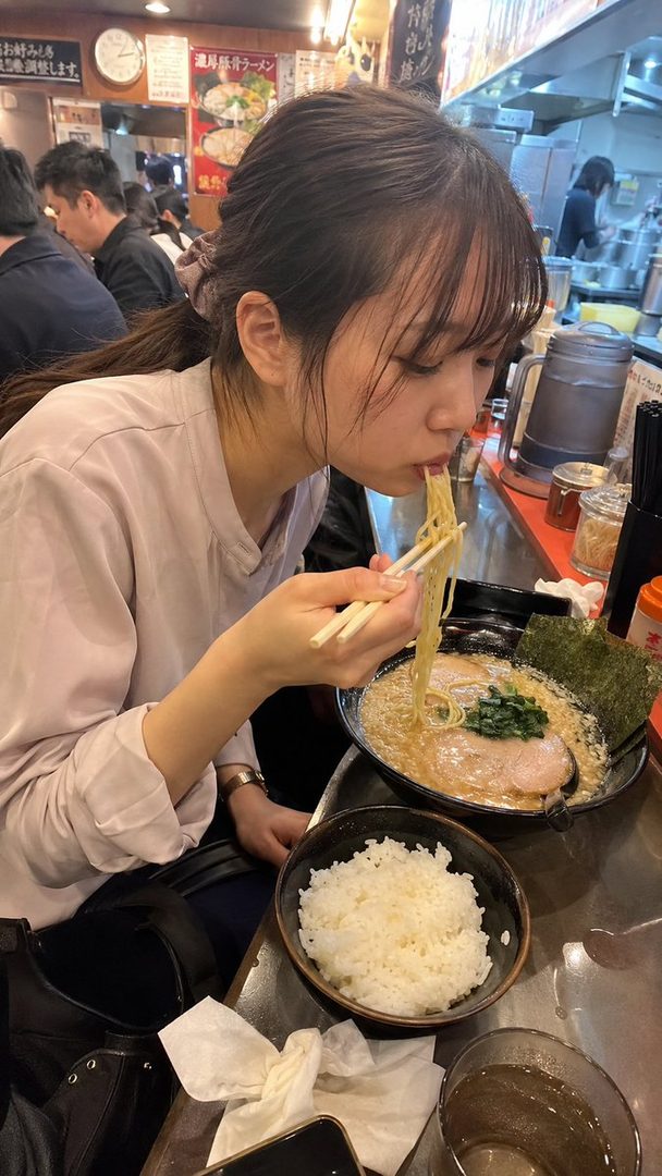 A young woman with dark brown hair tied in a ponytail is eating ramen with chopsticks at a busy ramen shop, with a bowl of rice next to her.