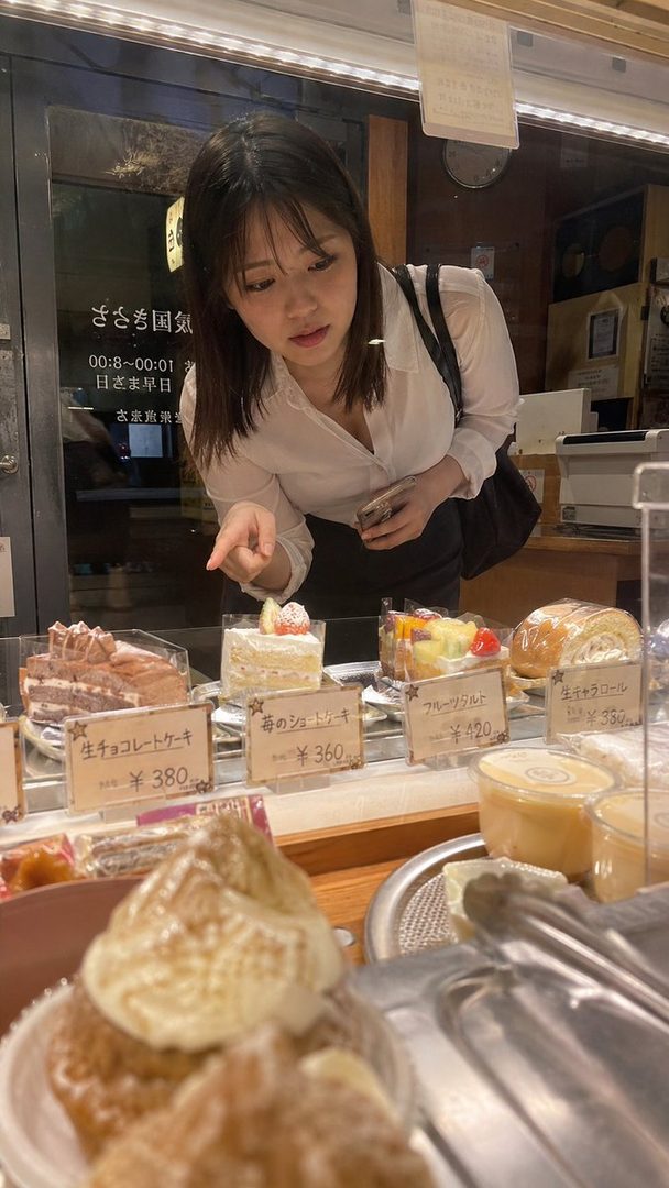 A young woman in a white shirt points at cakes in a bakery display case.