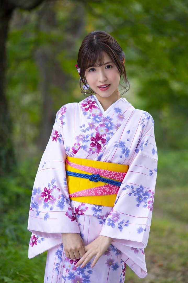 A young East Asian woman in a pink floral yukata with a yellow obi, posing outdoors with a gentle smile.