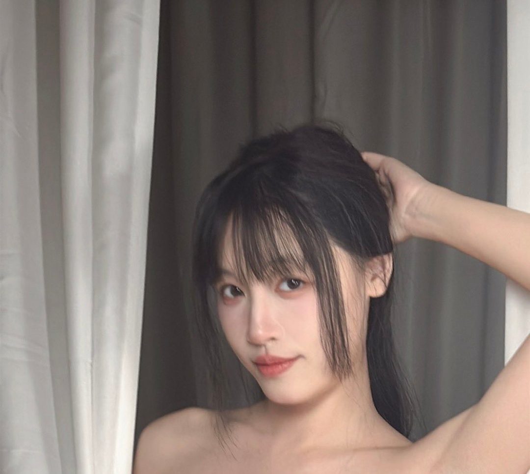 A close-up portrait of a young East Asian woman with black hair and bangs, looking playfully at the camera.