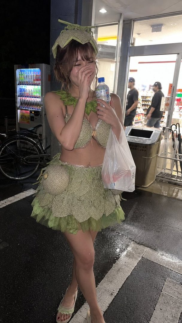 A woman in a playful melon-themed costume, including a bra top, mini skirt, and hat, laughs while holding a water bottle and plastic bag outside a convenience store at night.