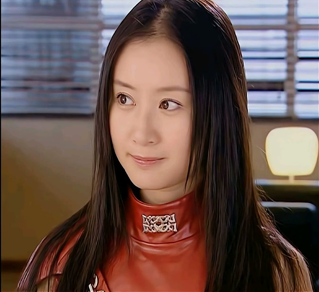 A young East Asian woman with long, dark hair, wearing a red high-necked outfit, looks to the side with a gentle smile.