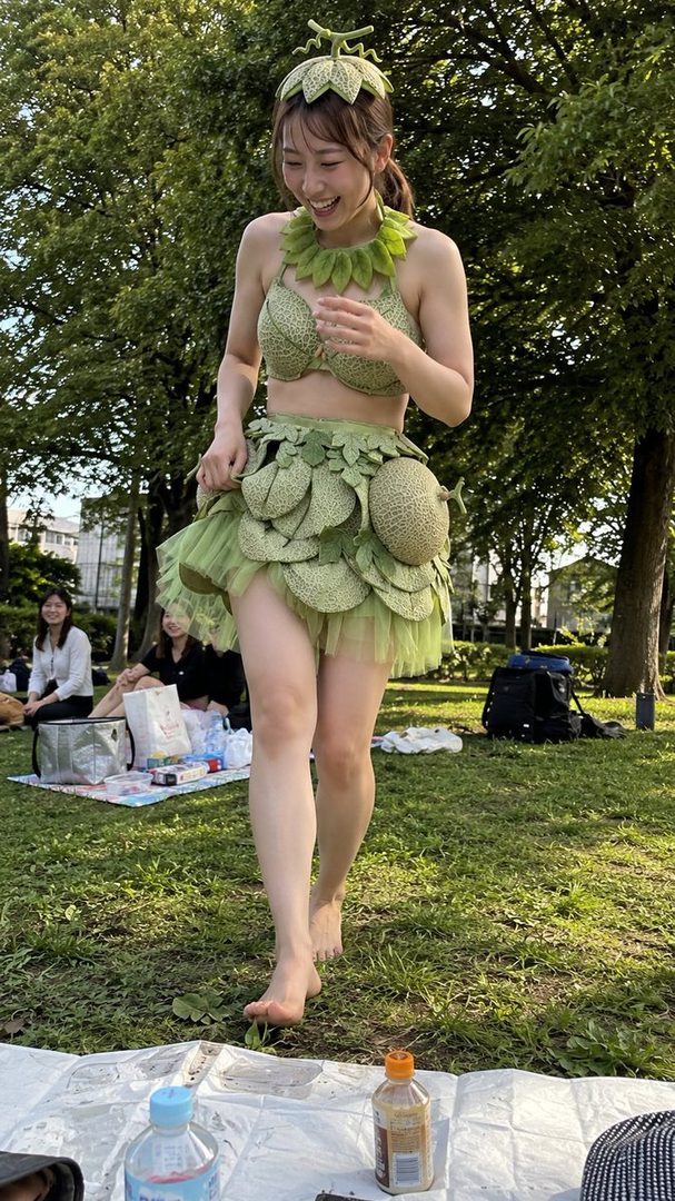 A woman in a green melon-themed costume, including a bikini top, tutu skirt, and hat, walks barefoot in a park and laughs.