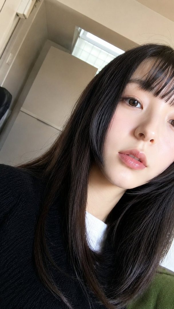 A close-up selfie of a young East Asian woman with long, dark hair and bangs, wearing a black sweater over a white shirt. She has a soft, cute expression and her lips are slightly parted.