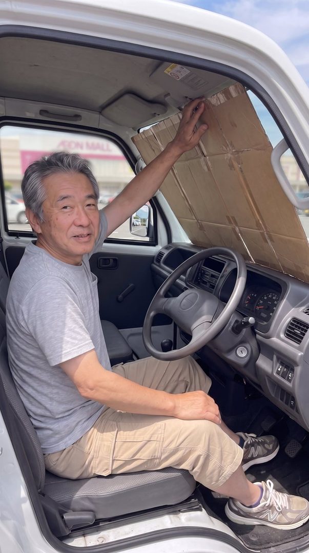 An elderly East Asian man sits in the driver's seat of a white van, adjusting a cardboard sunshade on the windshield.
