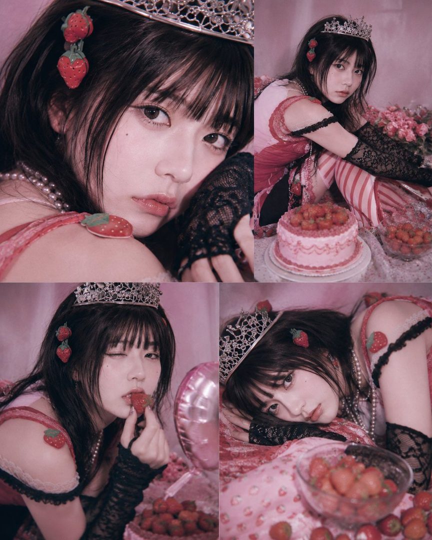 A young woman with dark hair, wearing a tiara and strawberry-themed accessories, poses playfully with strawberries and a cake.