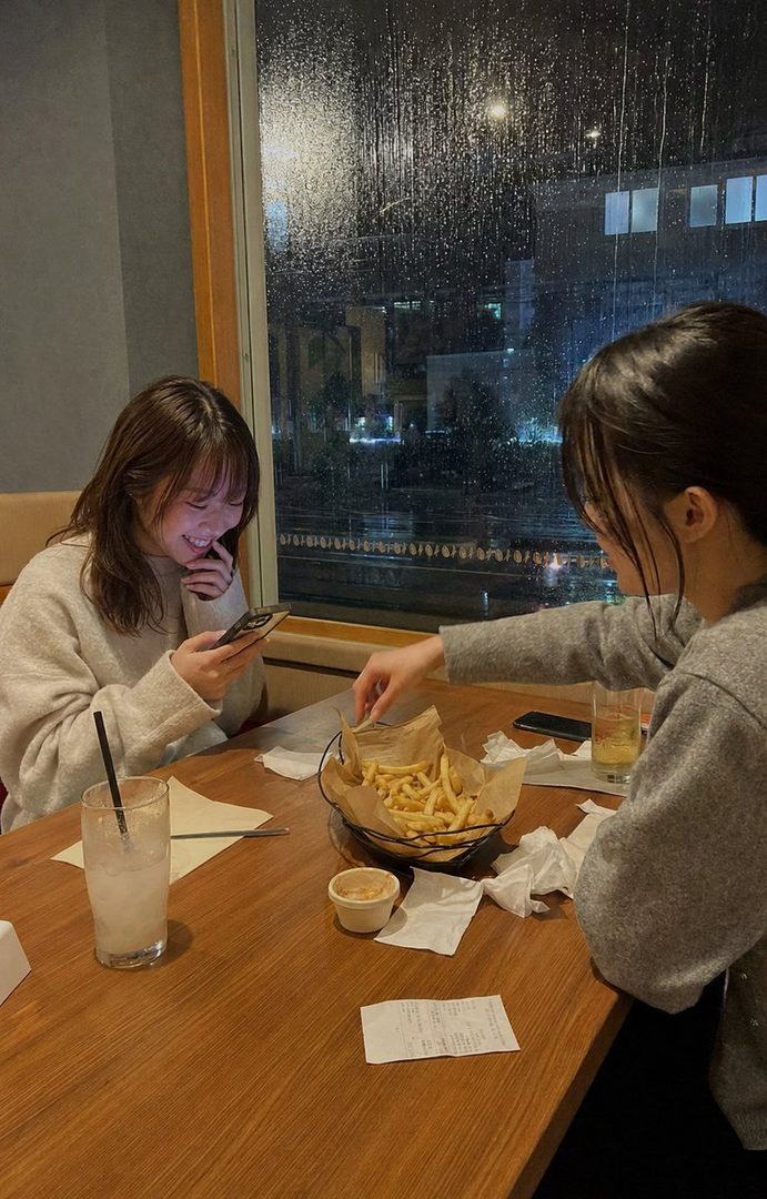 Two young women are enjoying french fries at a restaurant on a rainy night, with one smiling while looking at her phone.