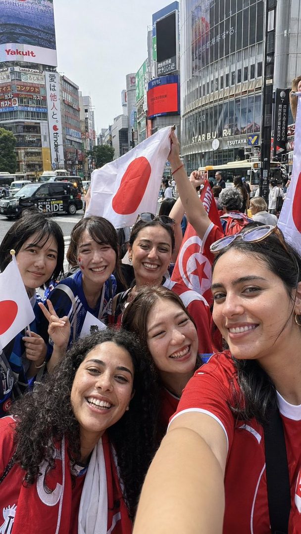 A group of diverse young women, some wearing red and blue sports jerseys and holding Japanese and Tunisian flags, are smiling and taking a selfie on a busy city street in Japan.
