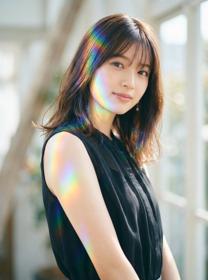 A young woman with long brown hair and bangs, wearing a black sleeveless top, is illuminated by a rainbow light effect on her face and arm. She looks towards the camera with a gentle expression.
