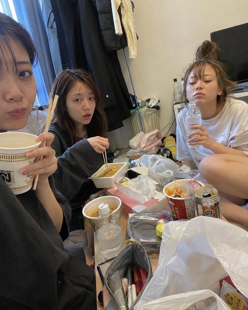 Three young women are casually eating instant noodles and drinking water on the floor of a messy room, looking tired but relaxed.