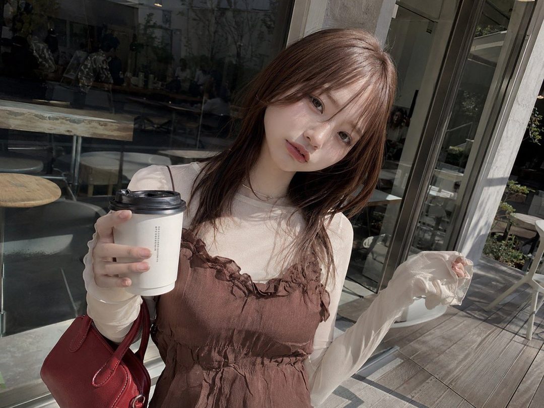A young East Asian woman with long brown hair holds a coffee cup and poses for a photo outdoors.