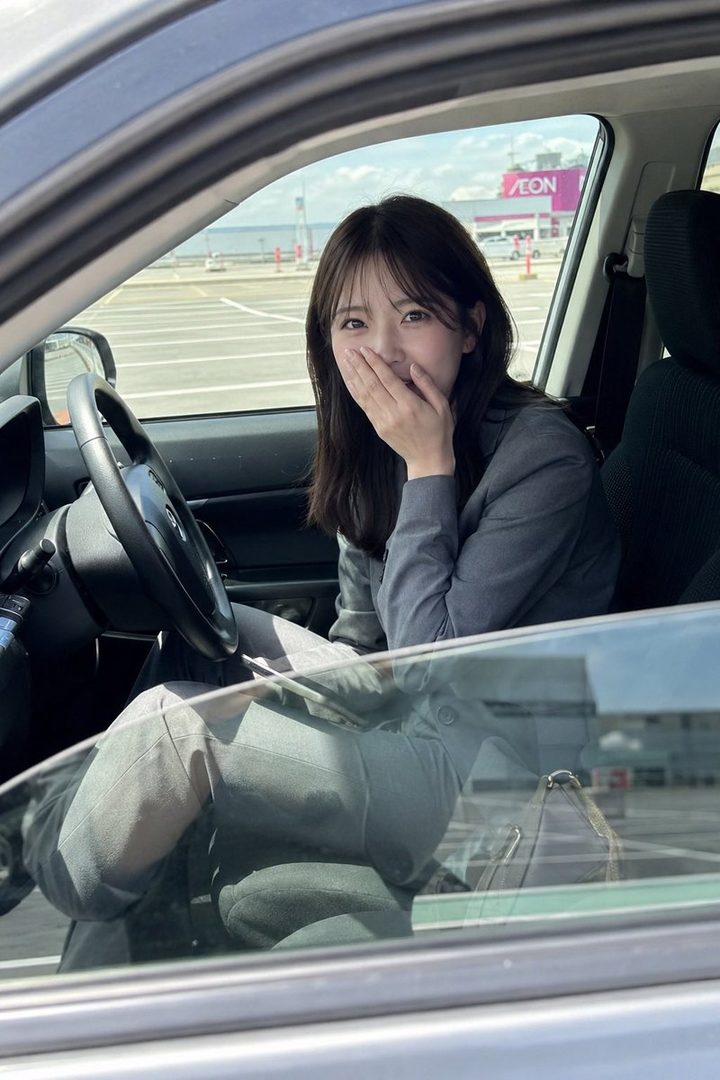 A young woman in a grey suit sits in the driver's seat of a car, covering her mouth with her hand and looking at the camera with a playful expression. She is in a parking lot with a shopping mall in the background.