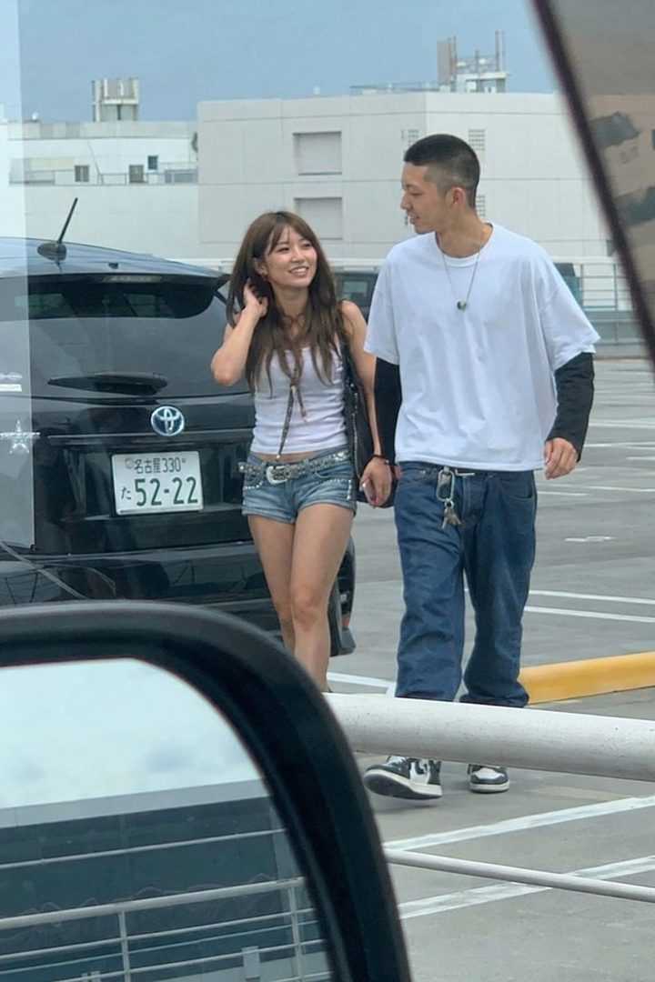 A candid shot of a young couple walking in a parking lot, with the woman smiling and wearing denim shorts and a white tank top, and the man in a white t-shirt and jeans.