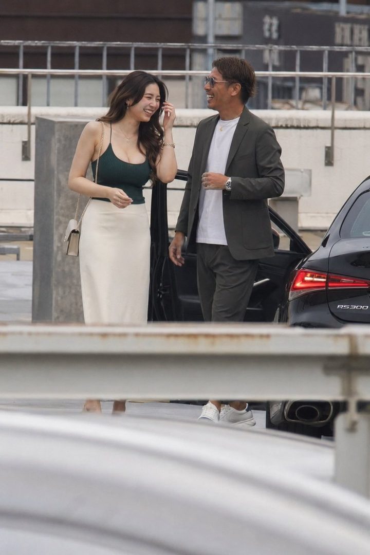 A smiling East Asian woman in a green tank top and white skirt stands next to a man in a suit, both appearing to be in a parking lot.