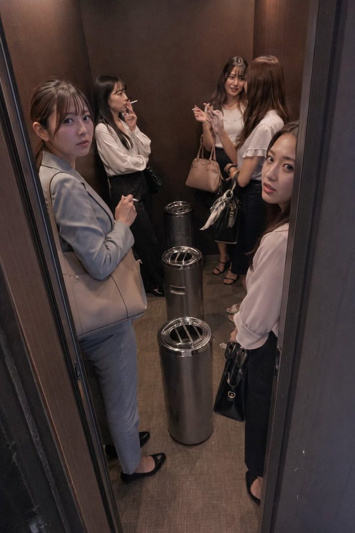 Four young women in business casual attire stand in a smoking area, two of them smoking cigarettes.