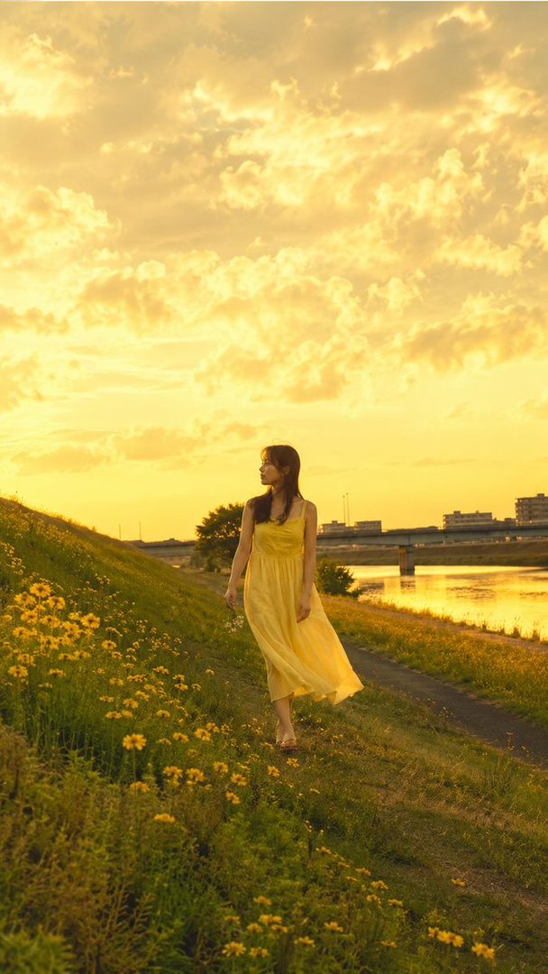 A woman in a flowing yellow dress walks barefoot on a grassy hill beside a river during a golden hour sunset.