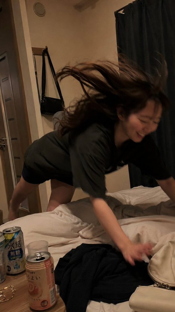 A young woman with long dark hair is playfully diving onto a bed in a dimly lit room, with beverage cans visible on a nearby table.