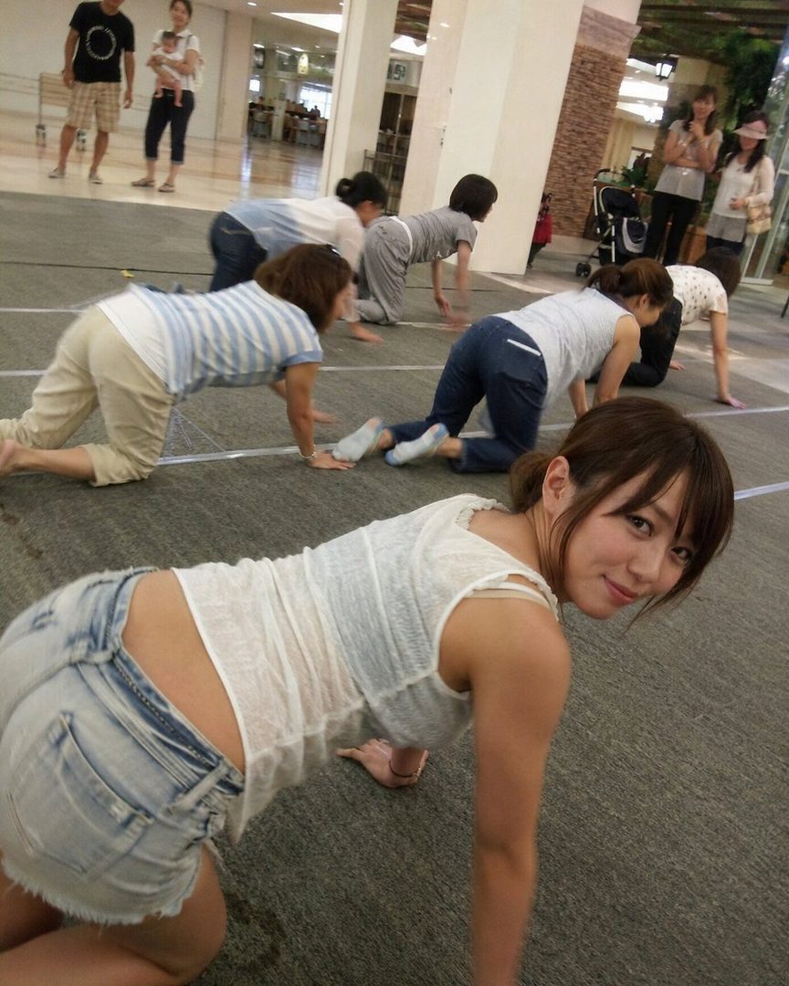 A young woman in denim shorts and a white top poses on her hands and knees, looking back at the camera with a playful smile. Other people are in the background in a similar pose.