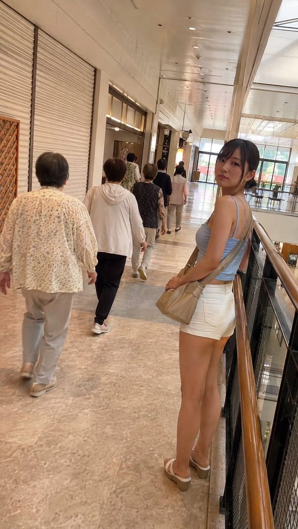 A young woman with black hair tied back, wearing a light blue tank top and white shorts, poses for a photo in a shopping mall hallway.