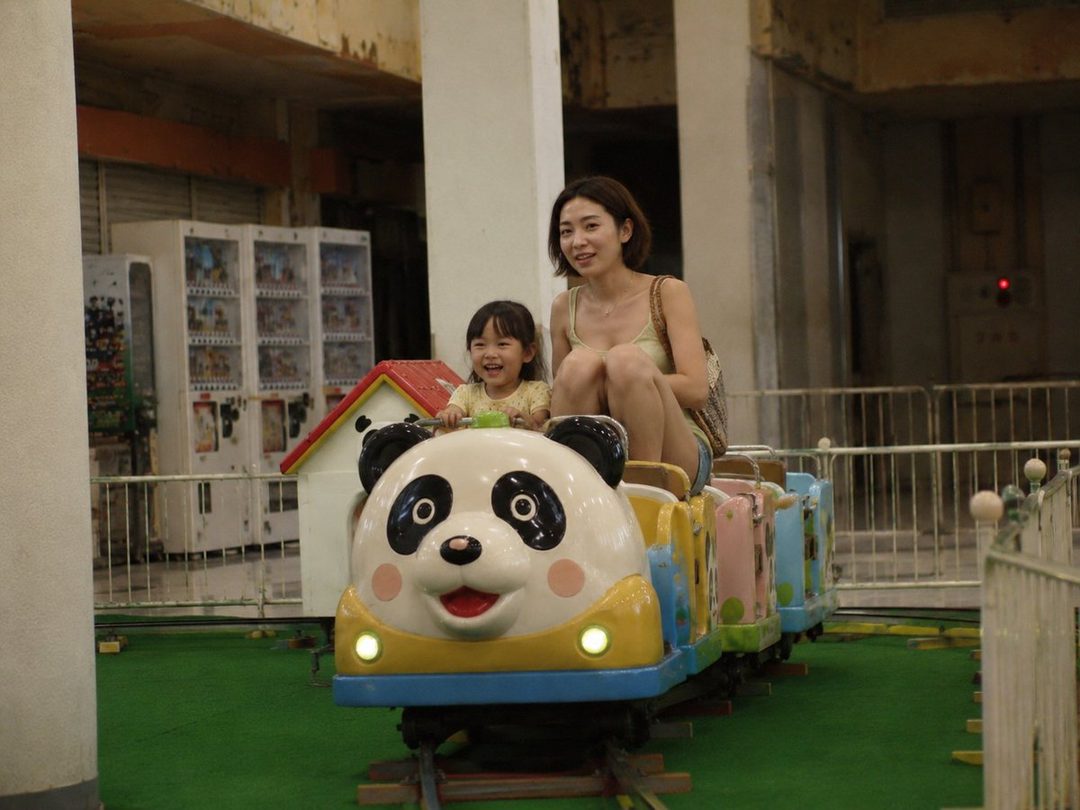 A mother and daughter ride a panda-themed train at an indoor amusement park.