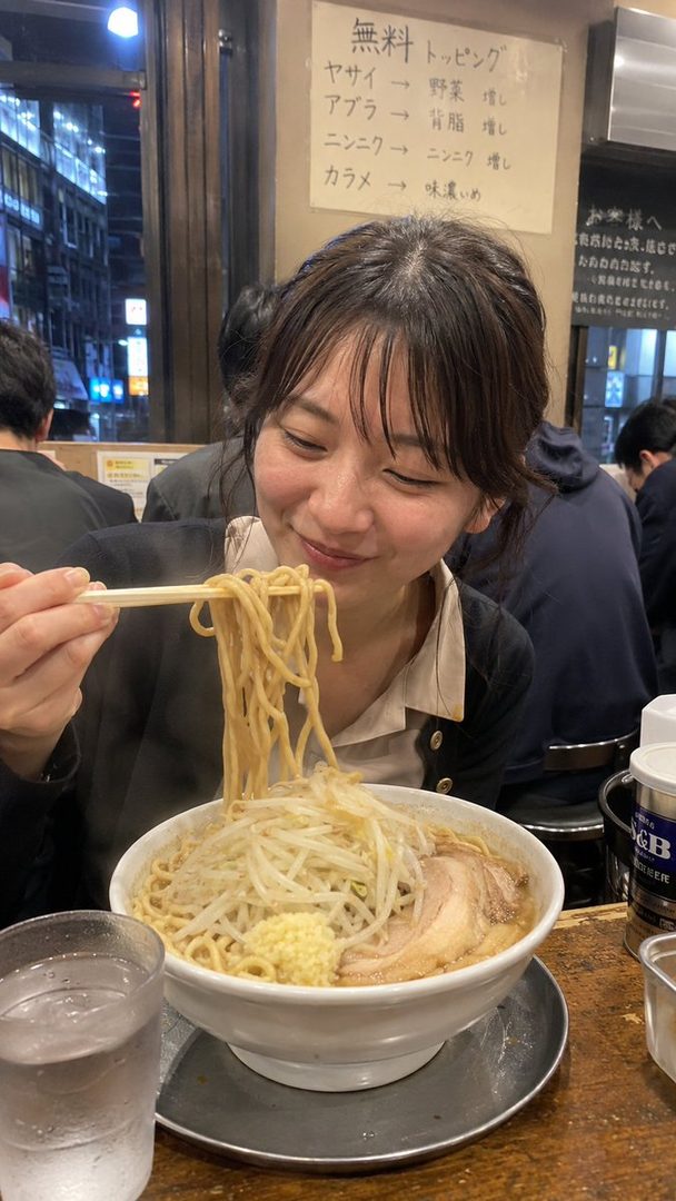 A young woman with dark hair and bangs smiles happily while eating a large bowl of ramen with chopsticks in a brightly lit ramen shop.