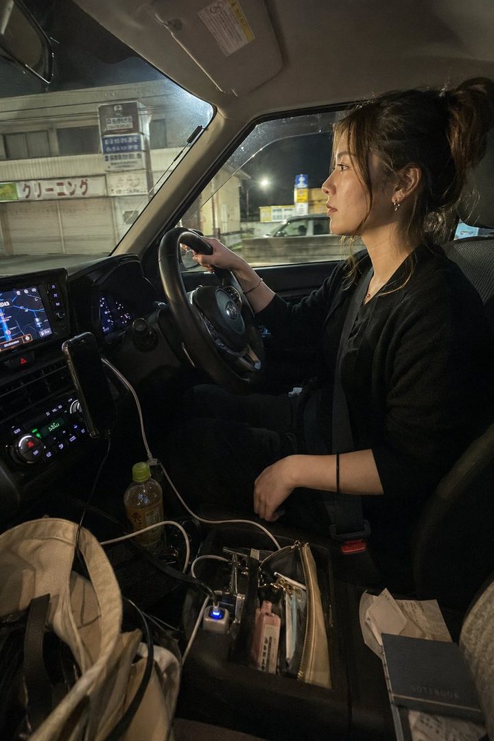 A woman with her hair in a messy bun is driving a car at night, looking out the window with a thoughtful expression.