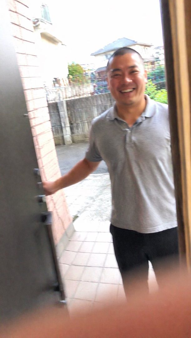 A smiling East Asian man in a grey polo shirt and black pants stands outside a doorway.