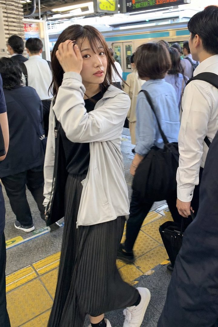 A young woman with dark brown hair and bangs, wearing a light gray jacket and a black pleated skirt, looks towards the camera while standing on a crowded train station platform.