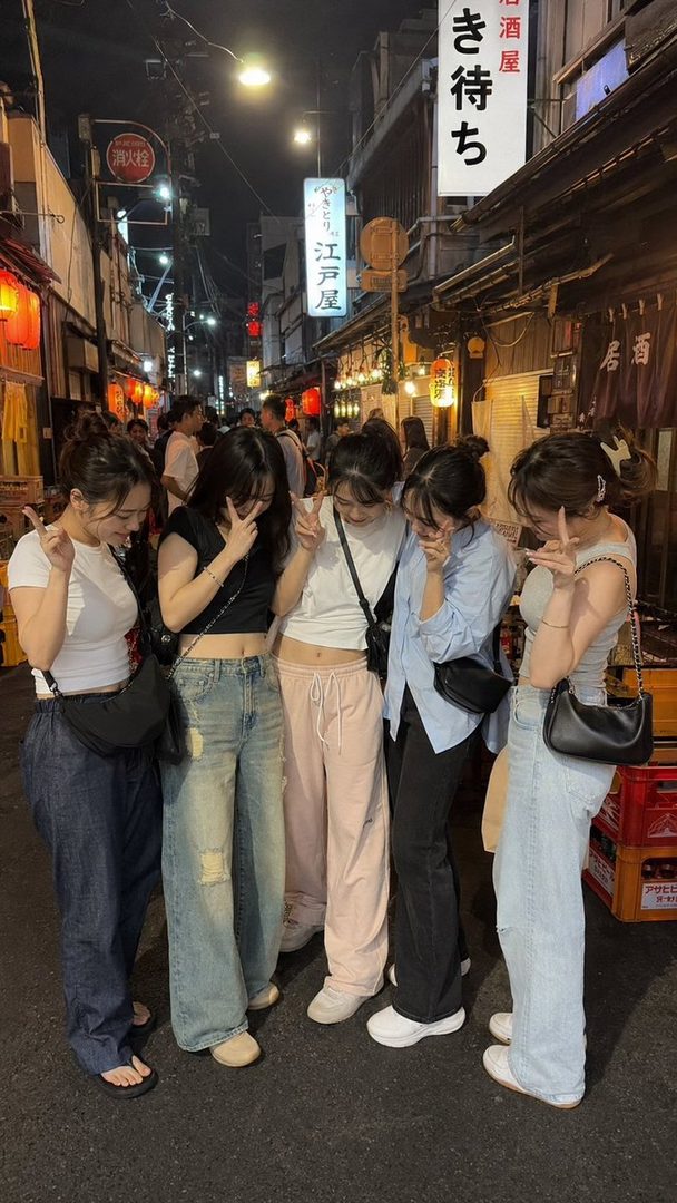 Five young women posing with peace signs on a lively street at night, with Japanese signs and lanterns in the background.