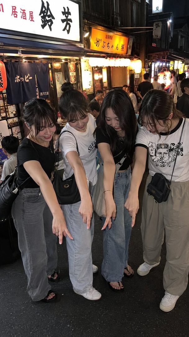 Four young women in casual attire are playfully pointing downwards on a lively street in Asakusa, Tokyo at night, surrounded by illuminated Japanese signs and lanterns.