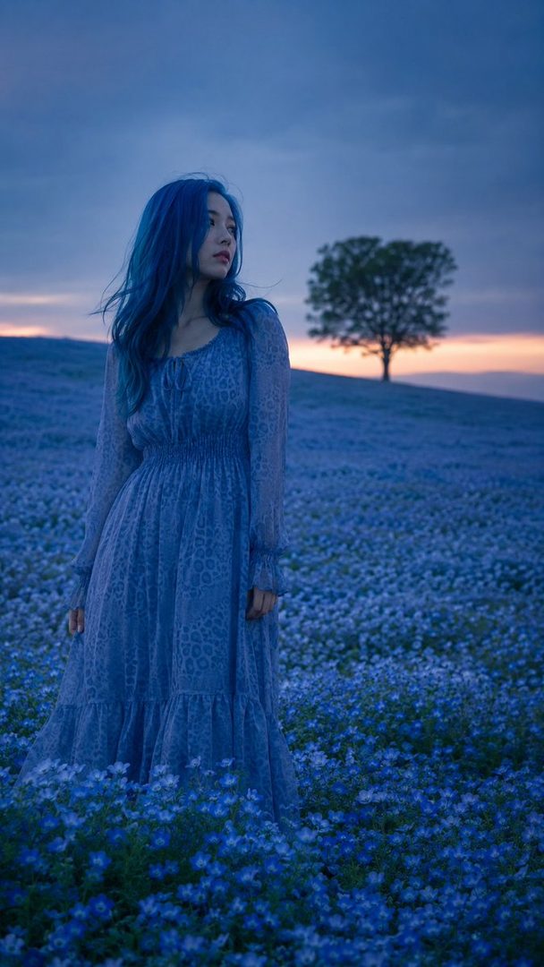 A woman with long blue hair and a blue dress stands in a field of blue nemophila flowers at dusk, with a single tree on the horizon.