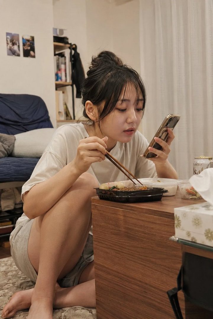 A young woman with dark hair in a messy bun sits on the floor, eating from a takeout container with chopsticks while looking at her phone in a cozy room.