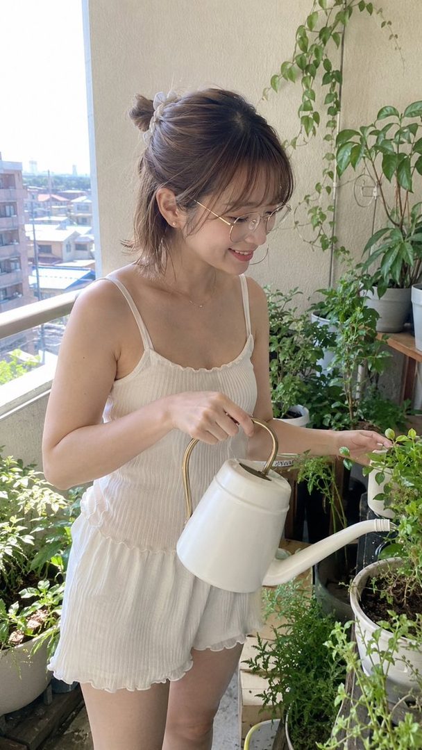 A young woman with short brown hair and glasses waters plants on a balcony.