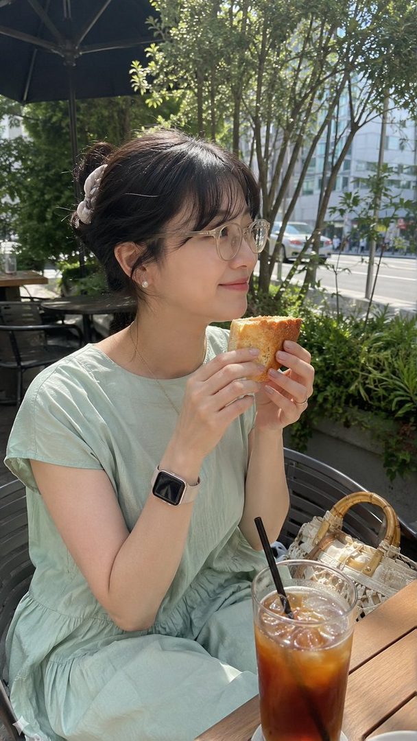 A young East Asian woman with dark hair tied up and wearing glasses, enjoying a sandwich and iced coffee at an outdoor cafe.
